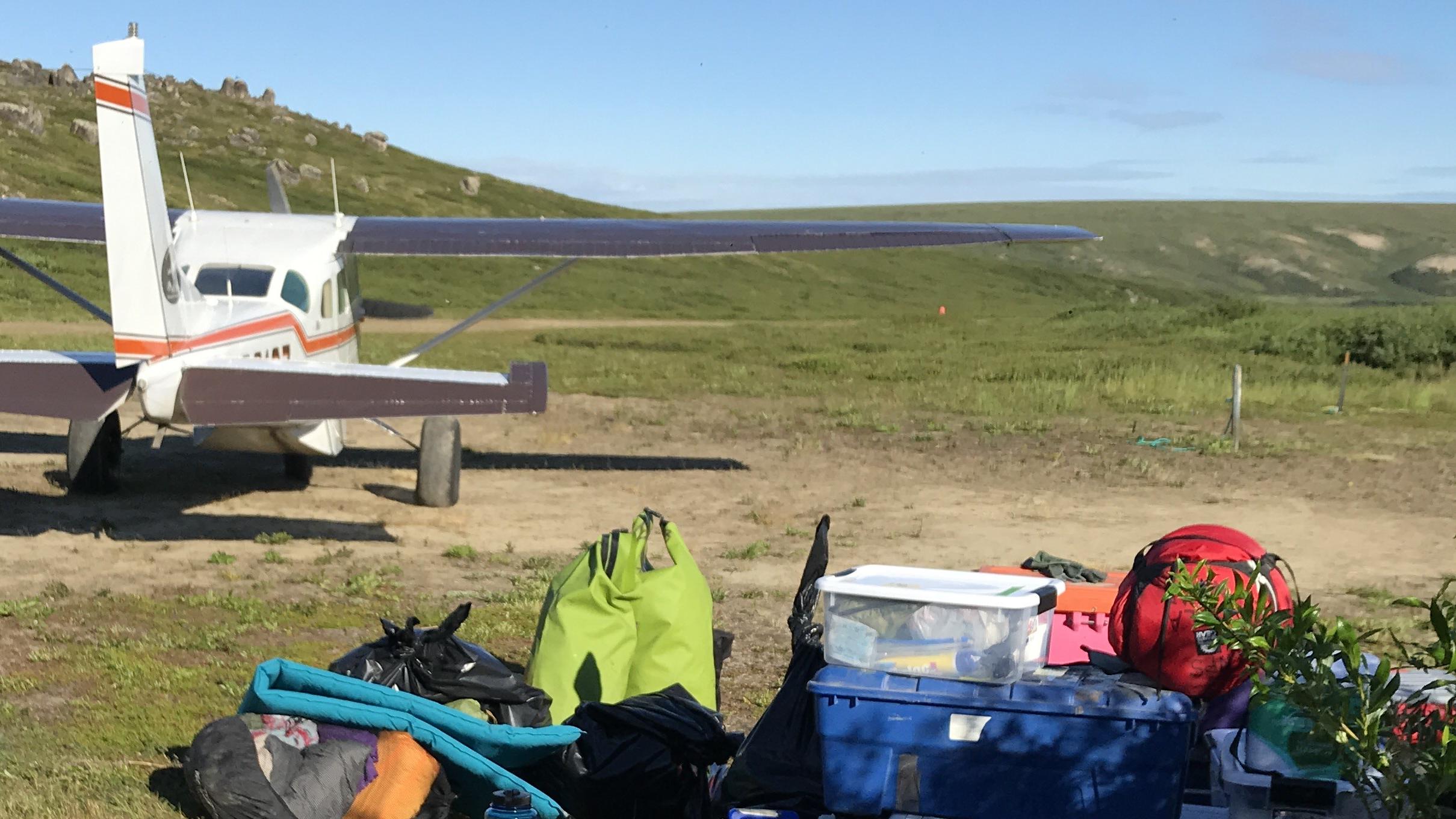Pile of gear in foreground and small plane in background in wilderness setting.