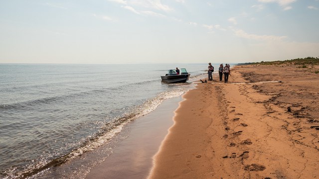 Photograph of a small boat on a sandy beach with people standing on the shoreline.