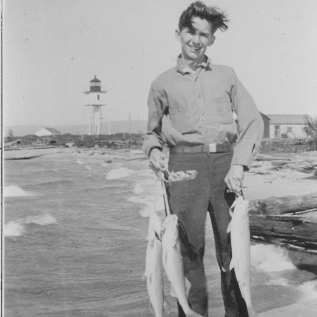 A young man standing on a pier holding three fish in front of a light station.