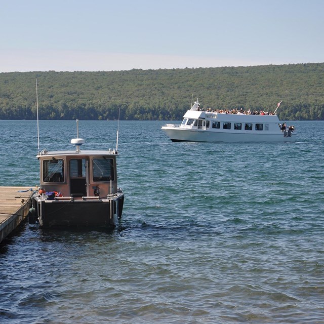 A small boat is tied to a dock, while a larger tour boat passes by.