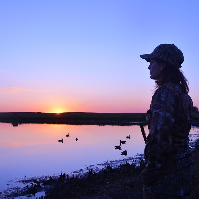 Silhouette of a hunter in front of pink and purple water and sky with a setting sun.
