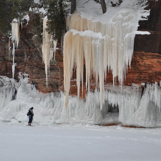 A lone person stands on a frozen lake looking at an ice-covered, sandstone cliff. 