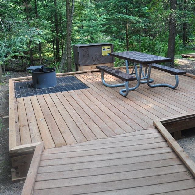 Raised wide boardwalk at campsite with accessible picnic table, bear box, and fire pit.