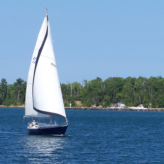 Sail boat with sails up on a body of water with land in background.