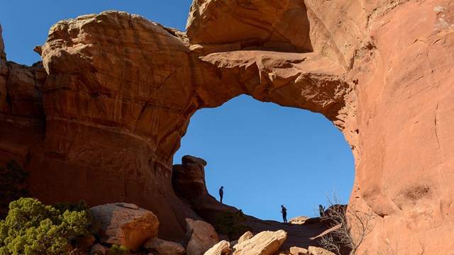 Two people standing underneath a sandstone arch silhouetted by the sun