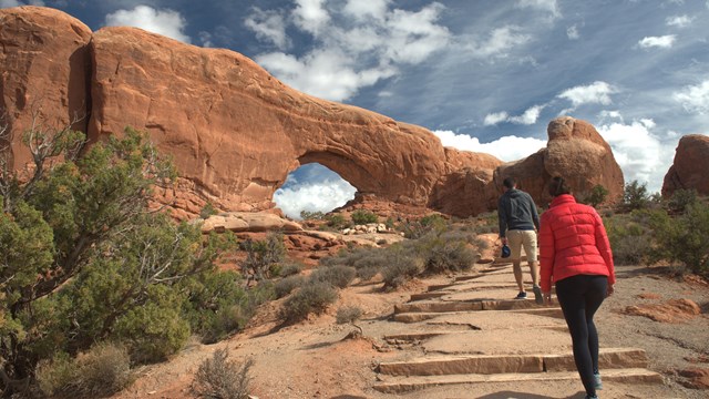 two hikers climb a trail past desert shrubs toward a large stone arch, blue sky and clouds above