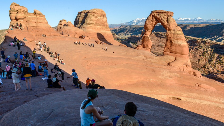 A crowd of people sit in a large rock basin, looking at a natural stone arch. 