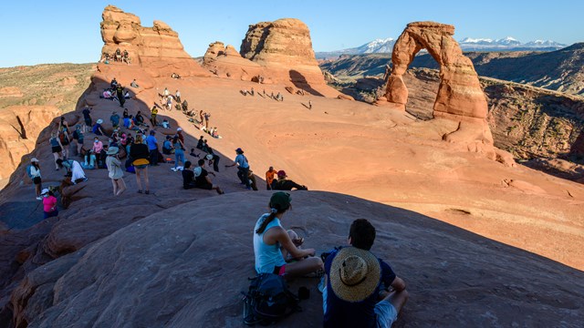 a crowd of people sit atop the edge of a sandstone bowl in the afternoon, gazing at delicate arch
