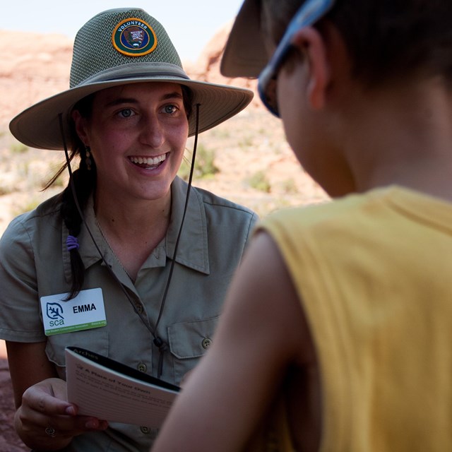 A ranger intern wearing a beige uniform shirt and hat talks to a child facing towards them