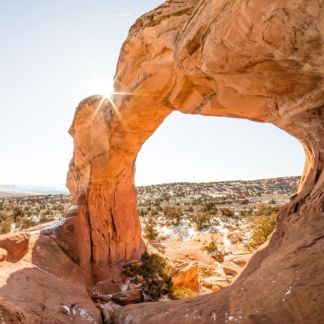 A red-orange colored rock arch with desert landscape behind it.