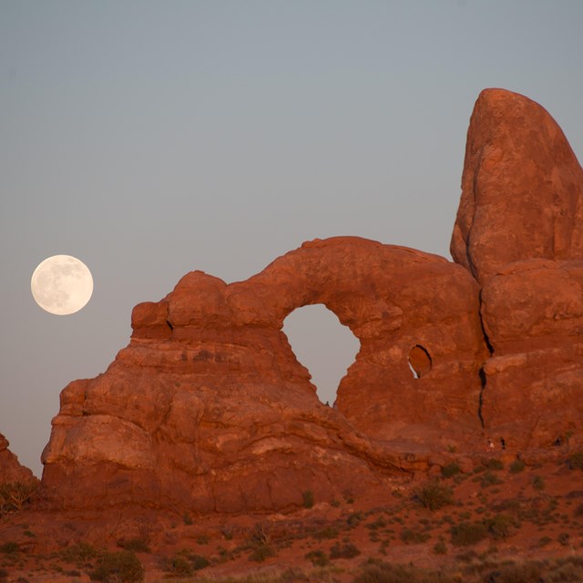 a large red rock arch with the rising full moon in the background