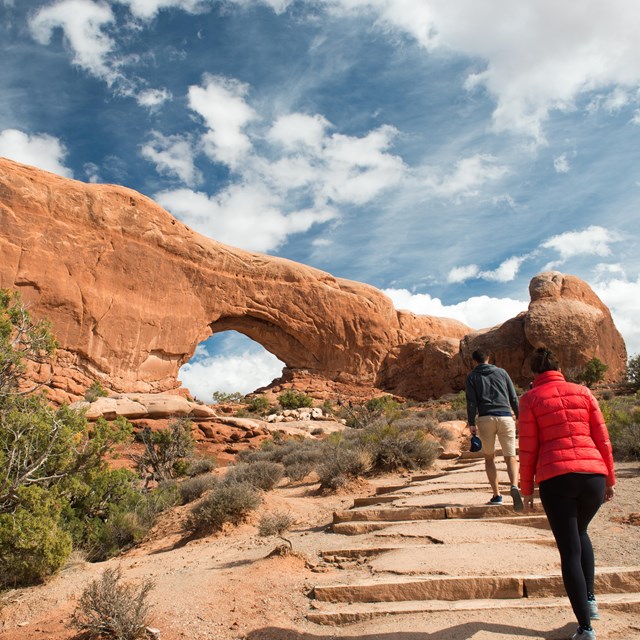 Two people walking up a set of steps on a trail leading to an arch.