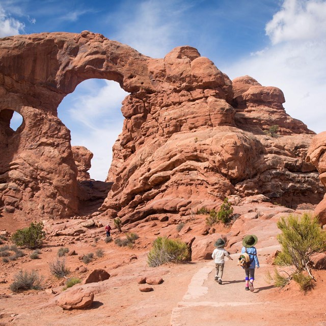 Children walk on a hard packed trail in front of a large red sandstone arch and canyon walls.
