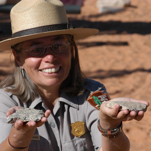 A woman in a park ranger uniform and straw hat hols up two gray rocks near her shoulders.