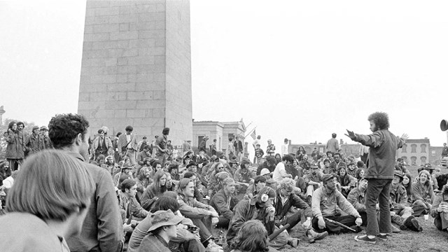 Black and white photo of a crowd standing and sitting at the base of the Bunker Hill Monument. 