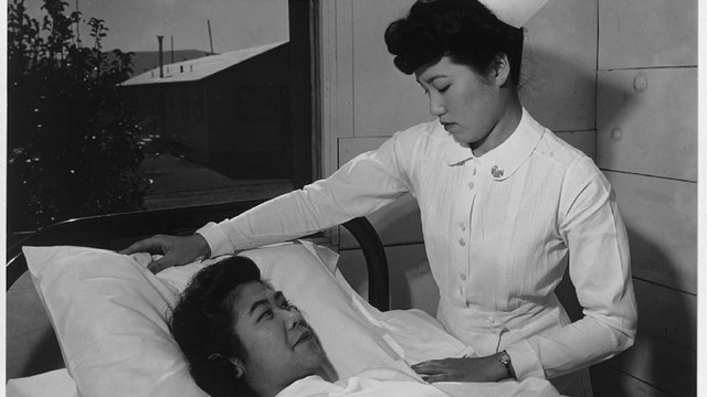 An East Asian woman in nurse's uniform tends to another East Asian woman in bed