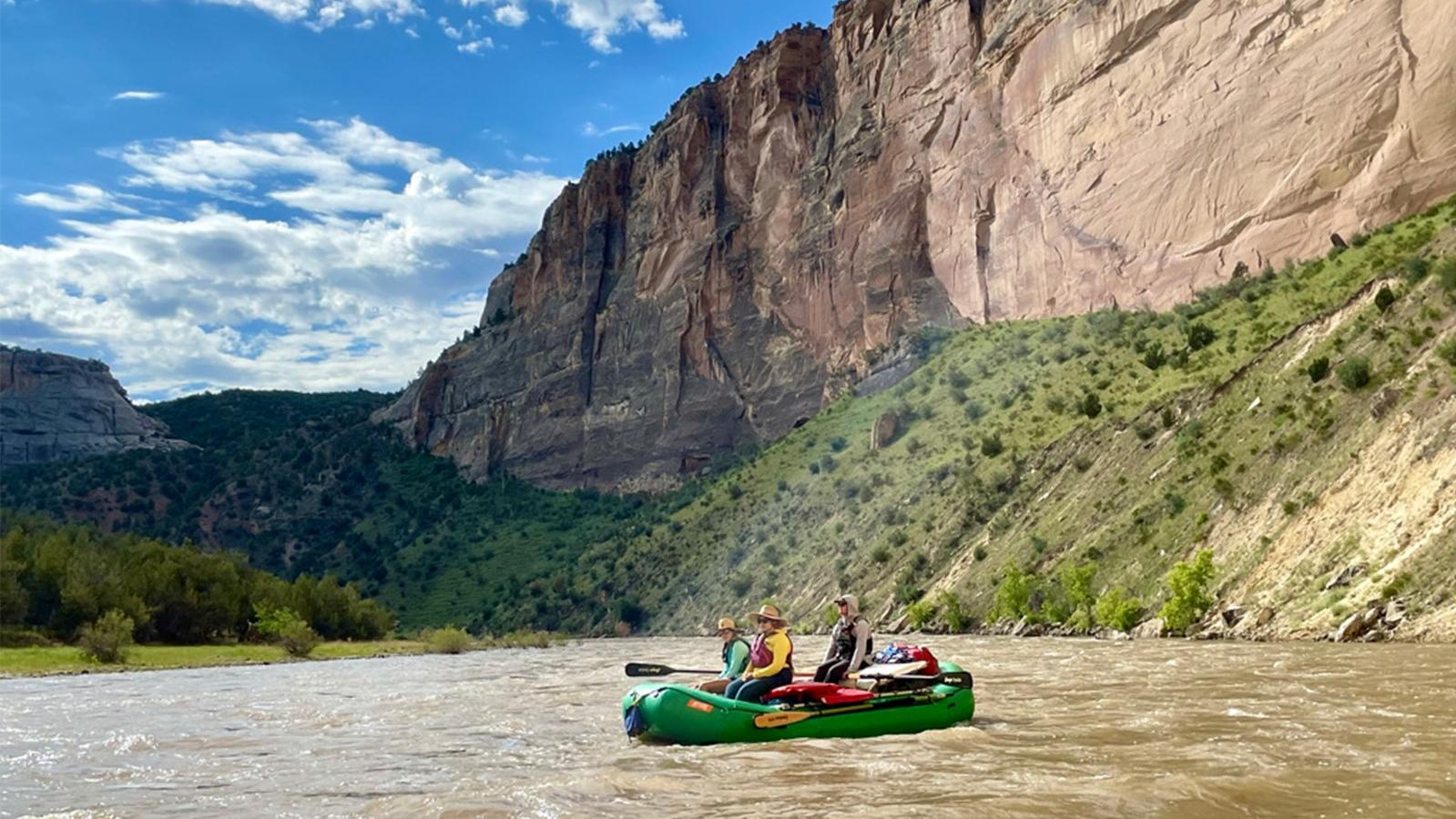 A green raft floats in a river. Canyon walls, green hill, blue sky, and clouds in background.
