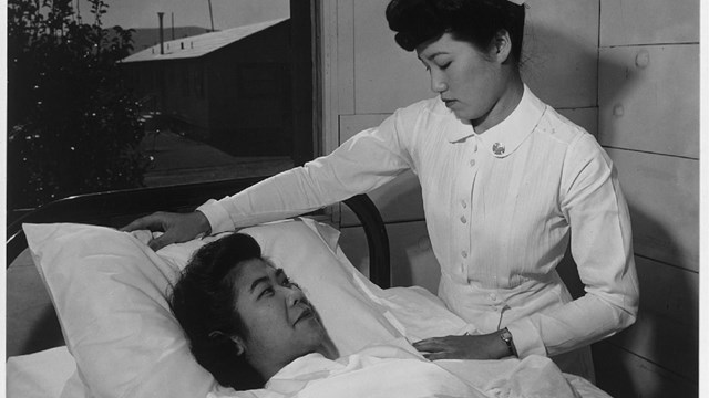 An East Asian woman in nurse's uniform adjusts pillow of another East Asian woman lying in bed