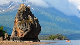 Image of a large monolith along a rocky lakeshore with mountains in the background.
