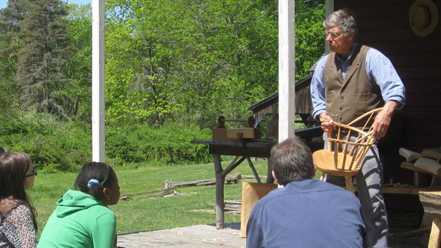 A man with a chair stands on a porch explaining the caining process to some teens.