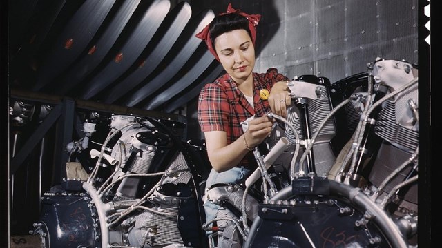 A white woman with brown hair wearing red shirt and red kerchief works on large mechanical device