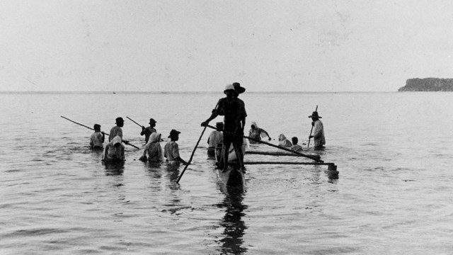 A man rows an outrigger canoe while a group of people fish in waist deep water around him.