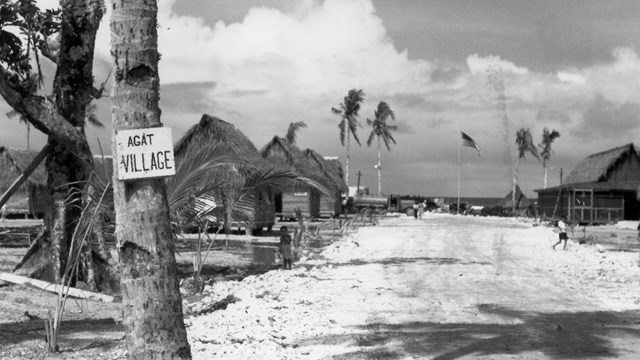 Dirt path lined with small wooden huts with palm front roofs. Sign nailed to tree says Agat Village