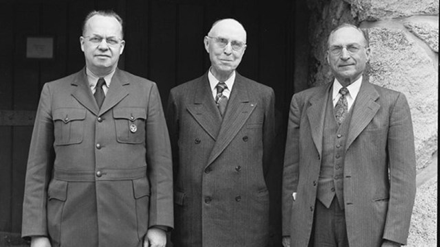Three white men in suits standing in front of a stone arch