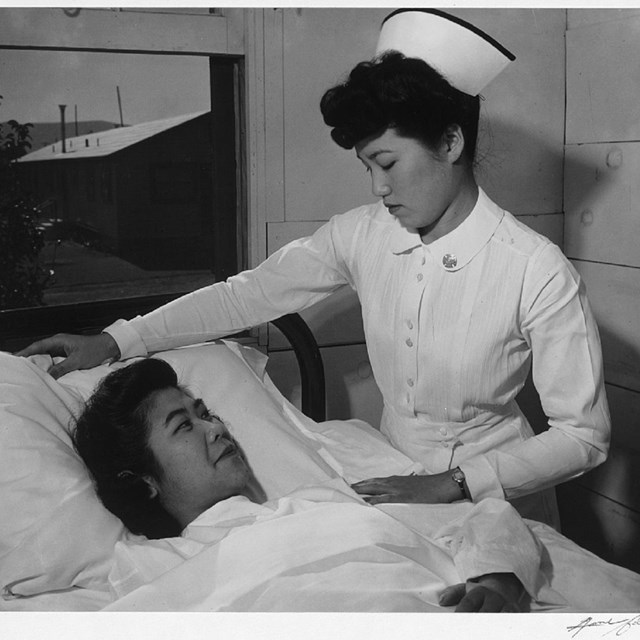 An East Asian woman in nurse's uniform tends to another East Asian woman lying in bed.