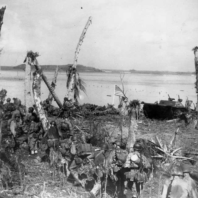 A line of soldiers march along a heavily bombed beach. A US flag flies next to an amtrac