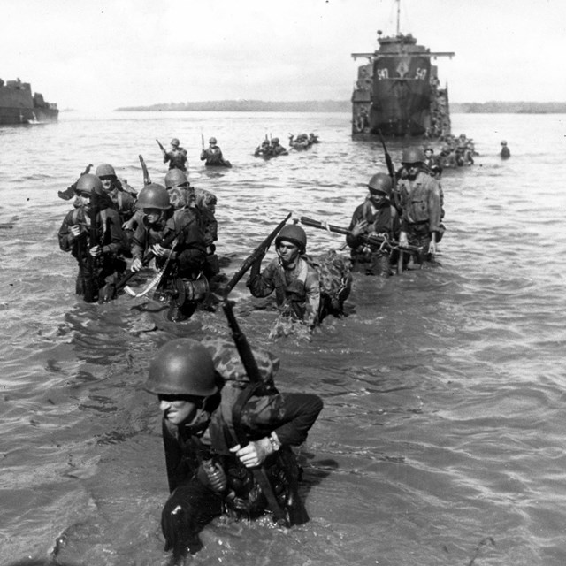 Uniformed soldiers carrying their packs and weapons wade through the water from an amtrac.