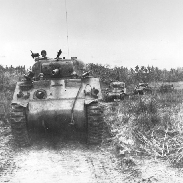 Three tanks drive along a dirt road. Three soldiers poke their heads out of the leading tank.