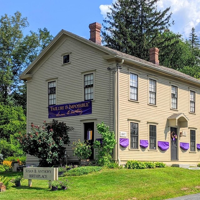 Large yellow two-story house with a sign in front saying Susan B. Anthony Birthplace