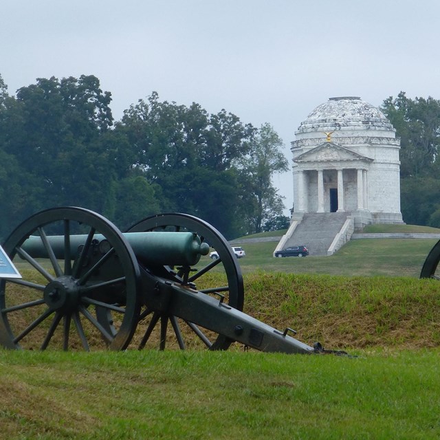 Two civil war cannons on grass battlefield. White building with columns and rounded roof set back