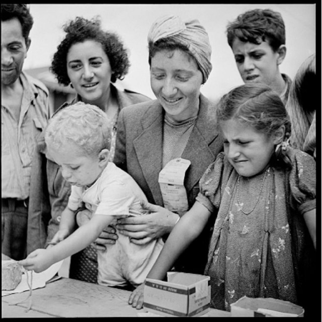 A group of men, women and children smile while leaning over papers on a table