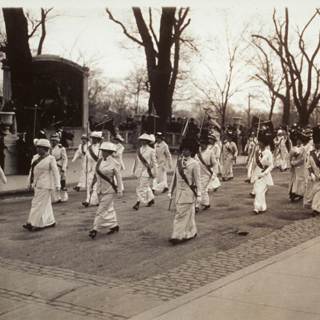 rows of women in white wearing sashes marching in a parade in front of the Shaw Memorial.