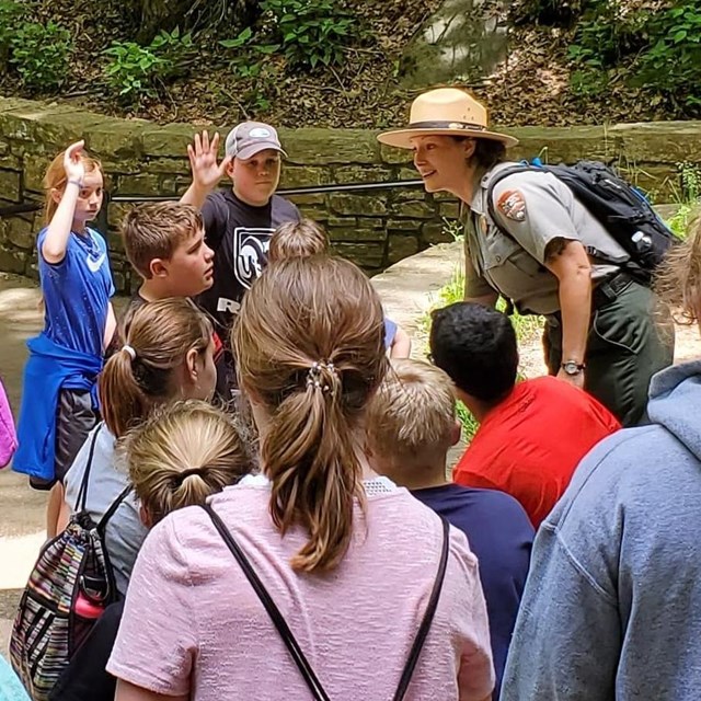 A woman in a green and gray NPS Uniform stands in front of a large group of students.