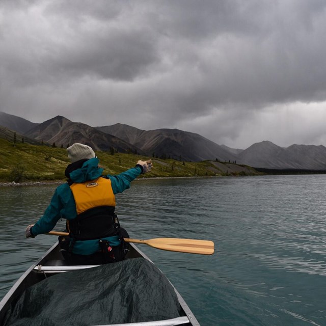 A woman in a canoe points towards mountains with storm clouds overhead