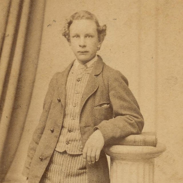 A photograph of a young Ernest Longfellow, leaning against a column and wearing a tailored jacket