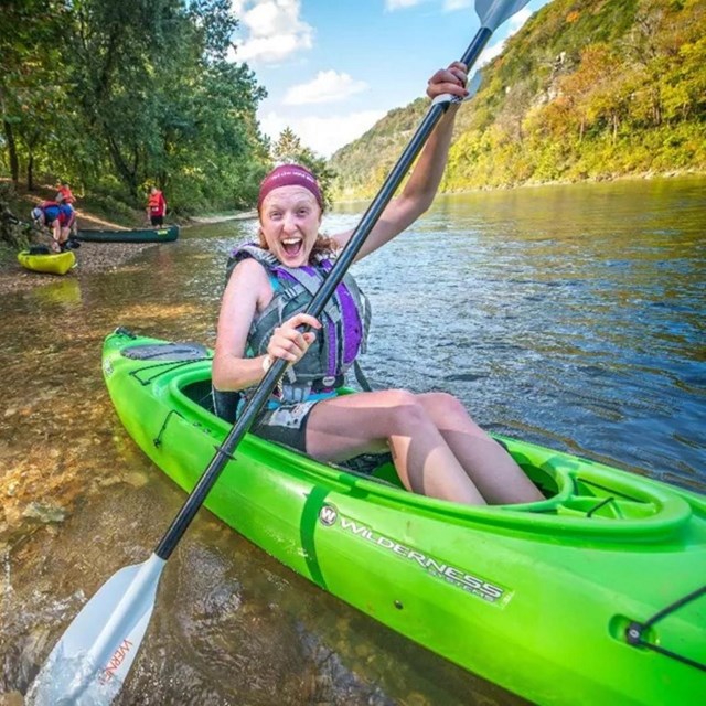 A woman smiling in a green kayak at the edge of a river.