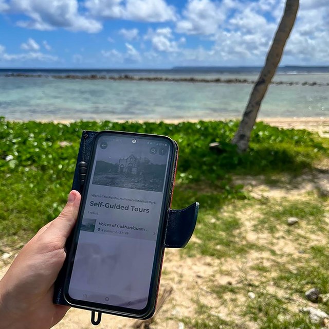 A cell phone open to the self-guided tours page of WAPA's NPS app. In the background is the beach.