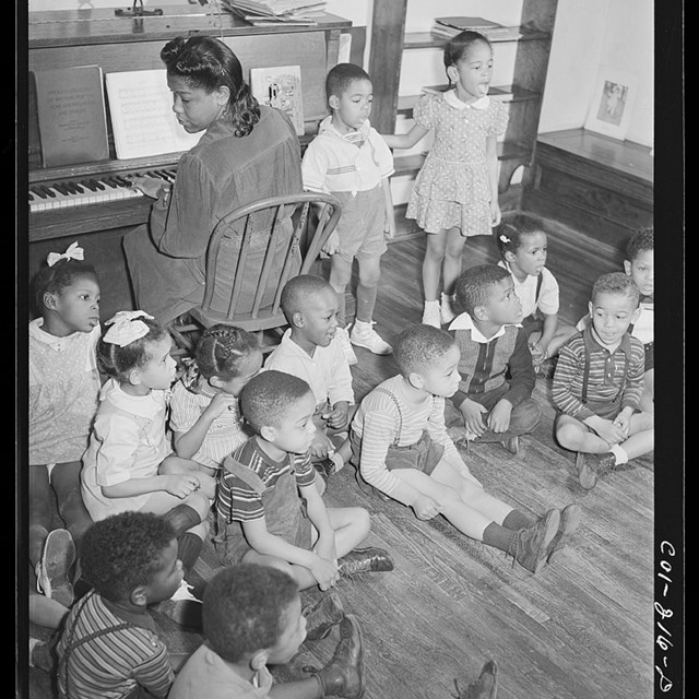 African American woman plays piano while several children sit and stand around her