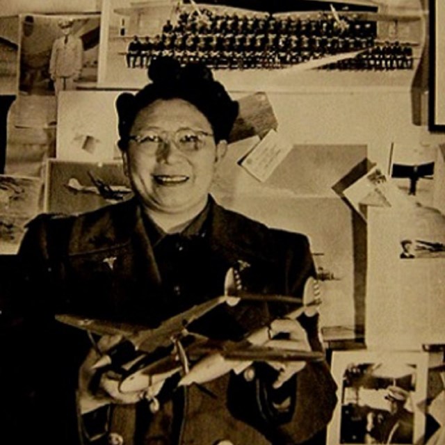 A middle aged East Asian woman in glasses smiles while holding a model airplane