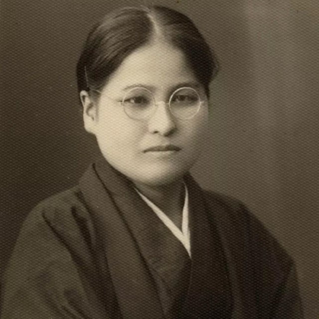 Forward facing photograph of a woman of East Asian descent wearing glasses & robes with slight smile