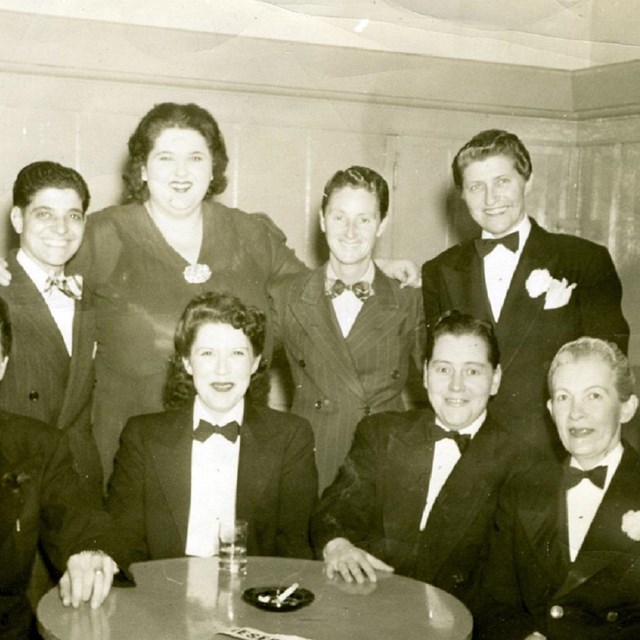 A group of people, most in suits and bow ties, hug and smile at the camera