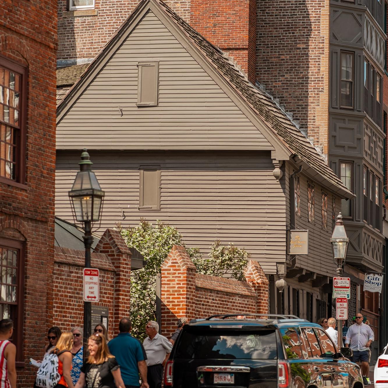 A photo of an old wooden home nestled in between red brick homes with cars and people out front