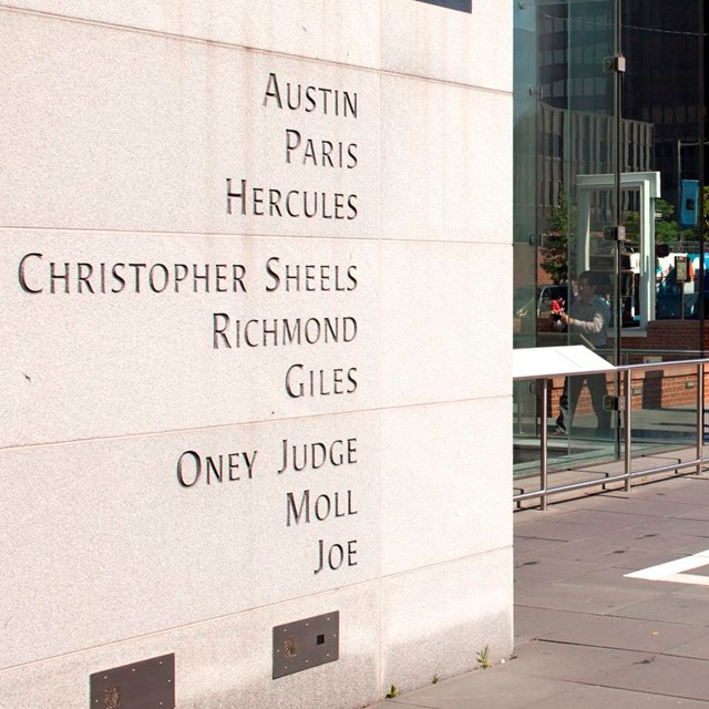 Photo of outdoor exhibit featuring a freestanding wall with the names of nine enslaved people.