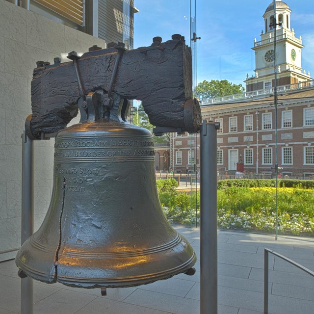 Photo of the Liberty Bell suspended on two steel posts with Independence Hall visible behind.