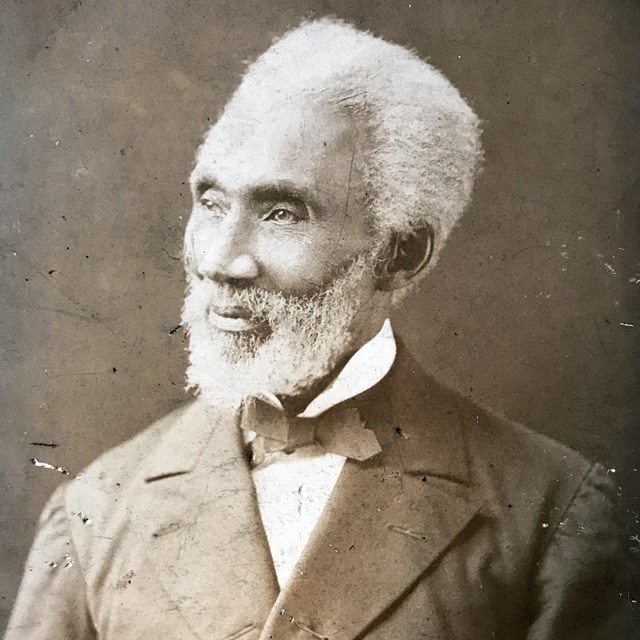 Historical black and white photo of a man with white hair and beard in a suit from the 1800s.