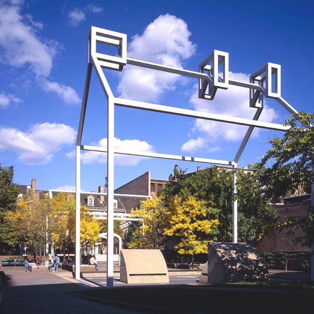 A white steel frame in the shape of a house towers over a courtyard.